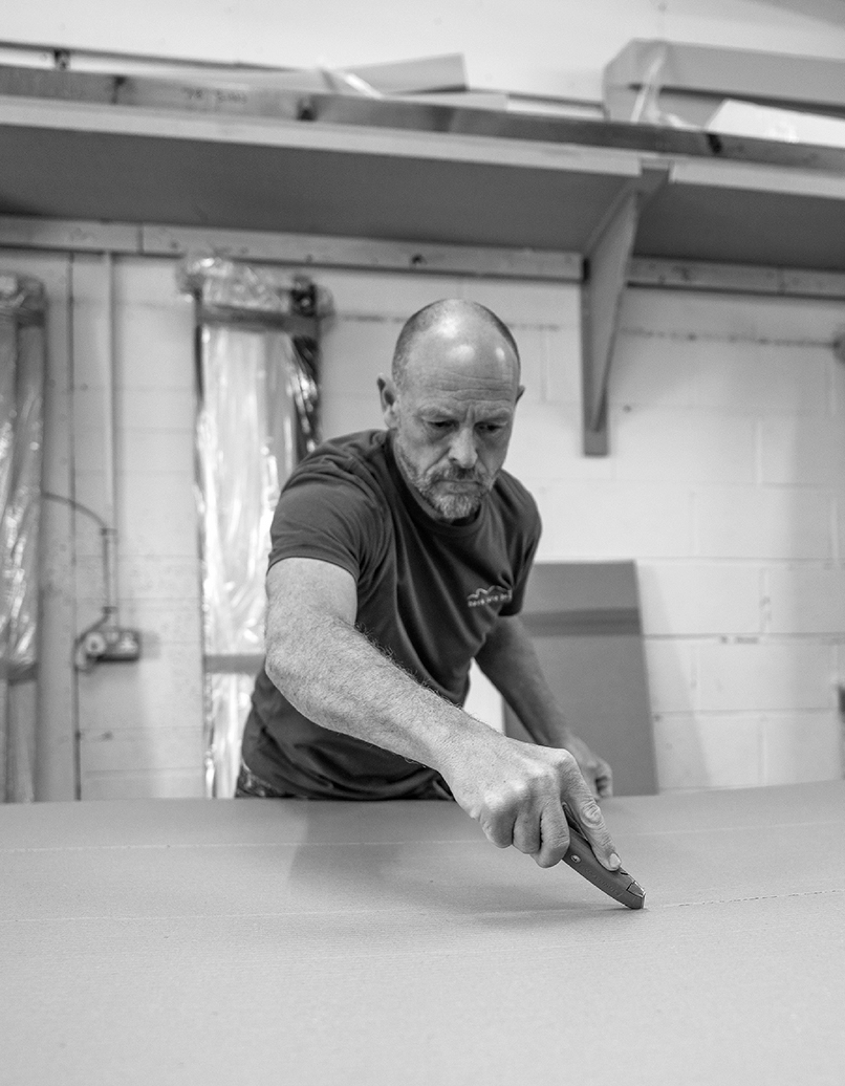A black and white photograph of a man in a workshop, wearing a dark t-shirt and using a knife to score or mark a large flat sheet of material. Shelves and industrial walls are visible in the background.