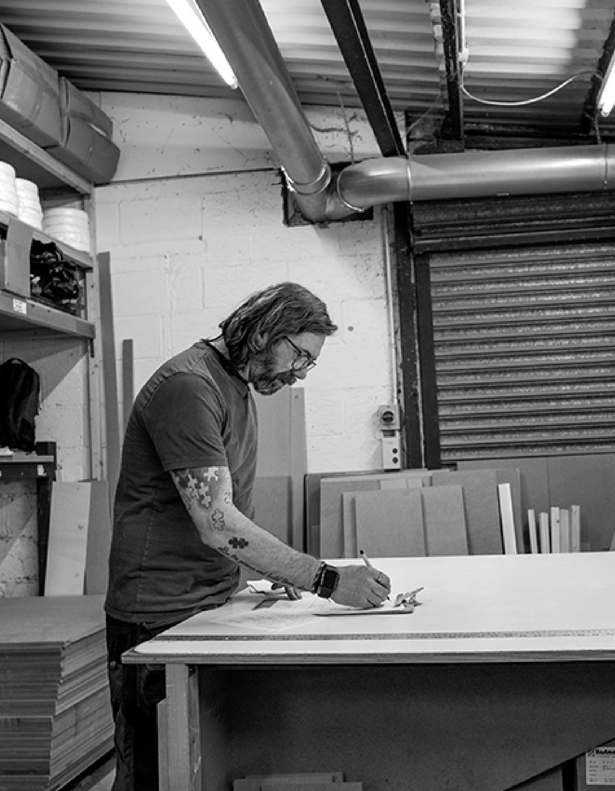A black and white photograph of a man with glasses and tattoos working at a drafting table in a workshop or studio, sketching on a large sheet of paper.