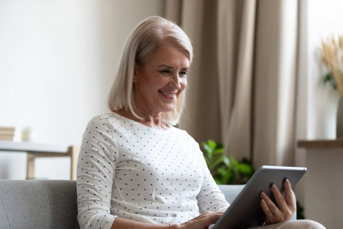An older woman with blonde hair sits on a gray sofa in a bright living room, smiling while holding a tablet computer.