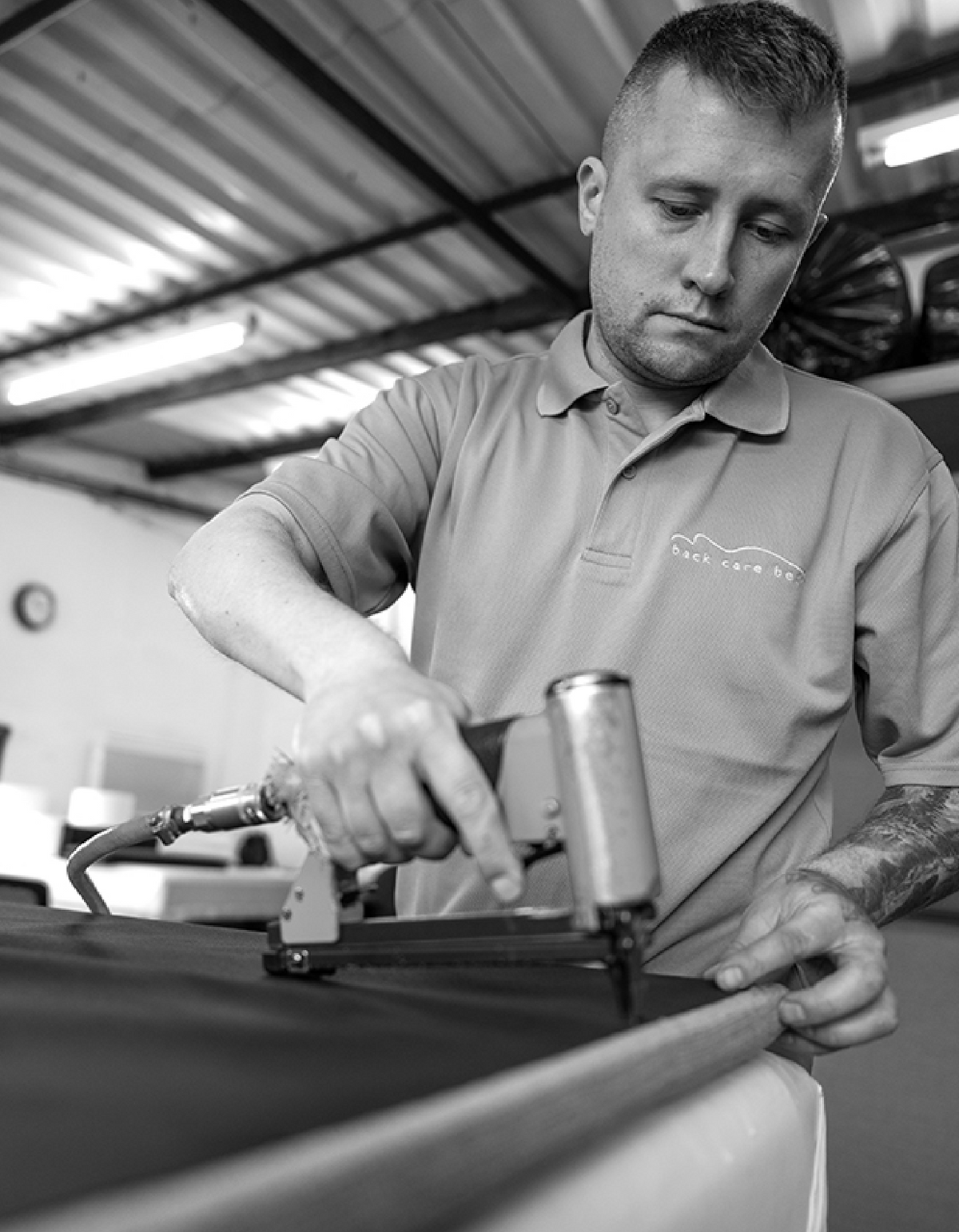 A man in a grey polo shirt with a 'Back Care Beds' logo is using a leather roller tool on a dark surface inside a workshop.